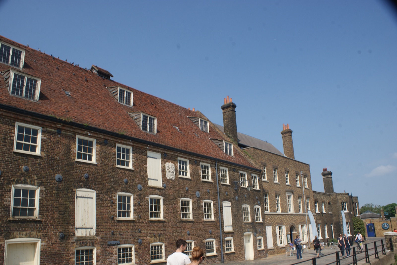 The long brick frontage of the mill on a sunny day, with visitors walking past and café tables outside.