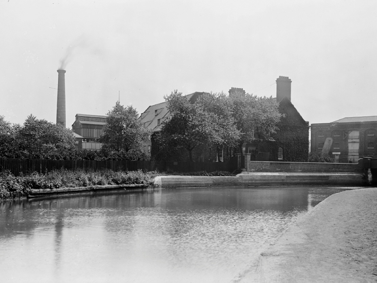 A historic photograph of the House Mill seen across the water, with its tall chimney and trees along the bank.